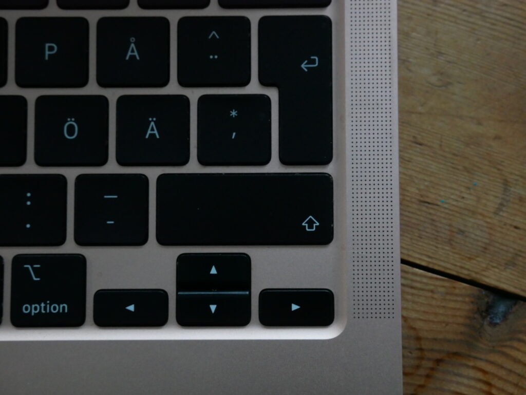 Nedre högra hörnet på ett Macbook, inzoomad något. Visar tangenter såsom Å, Ä och shift. Man ser lite av vad som ser ut att vara ett trägolv eller bord i bakgrunden.